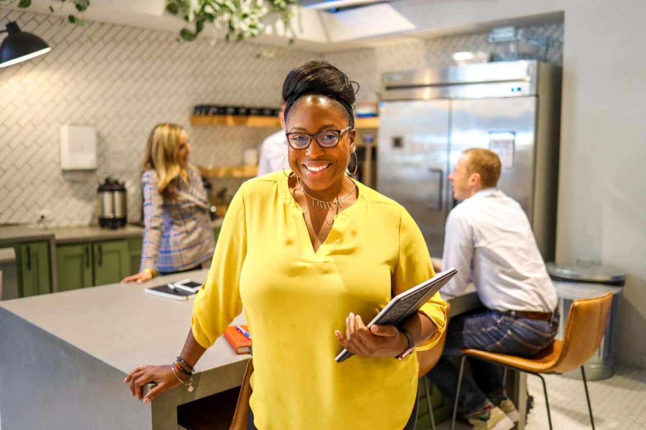 A professionally dressed, Pariveda leader standing in a kitchen with an Ipad in hand, smiling while a team of coworkers sit at a table in the background having a conversation.