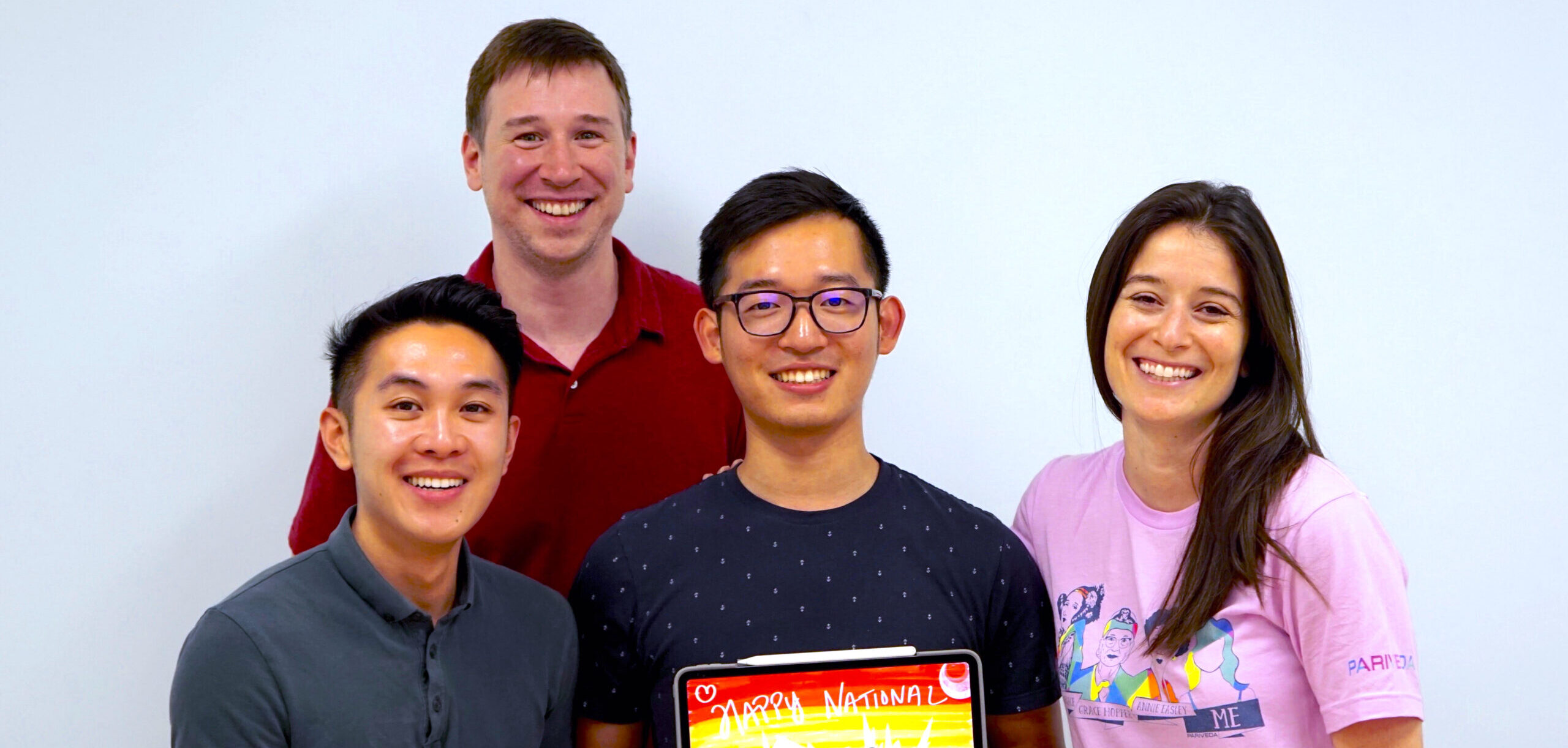 Pariveda employees stand in front of white background smiling and holding a sign stating "Happy National Coming Out Day"