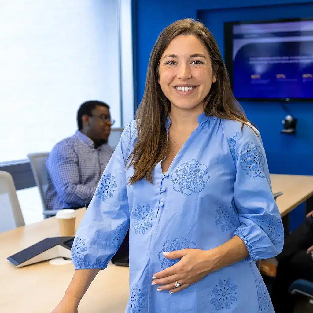 A pregnant woman in a blue embroidered dress stands at the head of a conference table, smiling confidently at the camera. Behind her, three colleagues, two men and one woman, are engaged in conversation in a modern office meeting room.