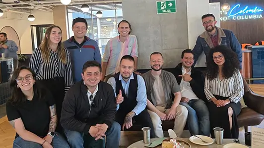 A group of eleven smiling people, casually dressed, gathered in a modern office lounge with coffee cups and pastries on the table. They are seated and standing together, posing for a photo under a sign that reads "It's Colombia, not Columbia," indicating the setting is Bogotá.
