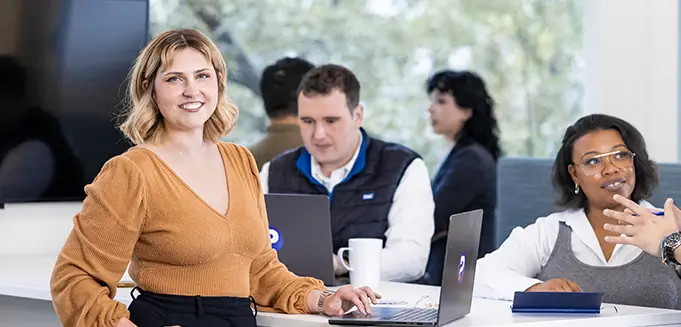 A group of professionals collaborating in a bright office space. A woman in a rust-colored blouse stands at the front smiling confidently, while two colleagues sit at the table with laptops open, engaged in conversation.