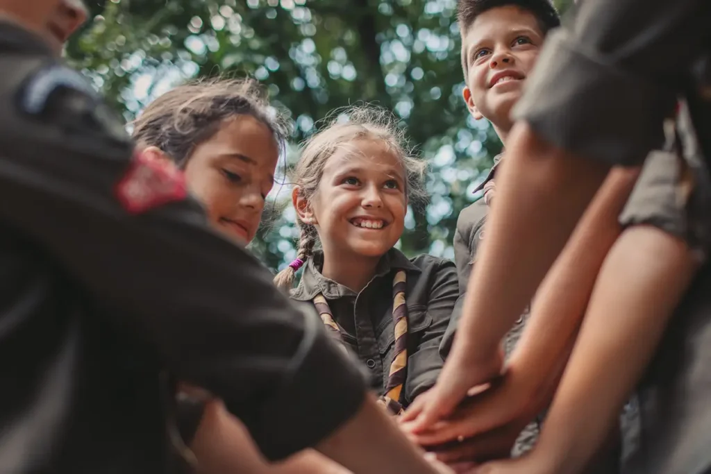 A group of smiling children in scout uniforms join hands in a team huddle outdoors, symbolizing unity, leadership, and collaboration.