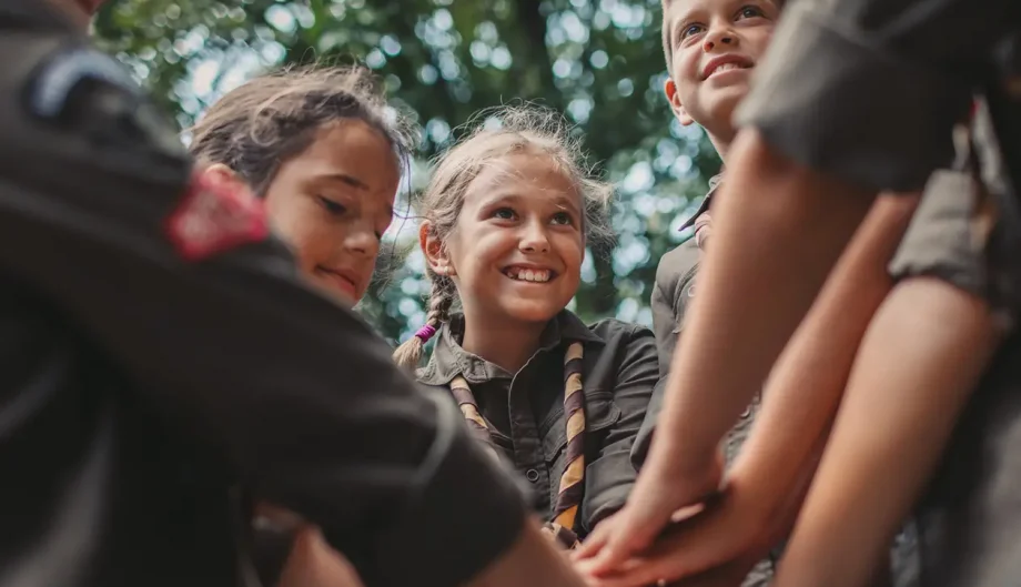 A group of smiling children in scout uniforms join hands in a team huddle outdoors, symbolizing unity, leadership, and collaboration.
