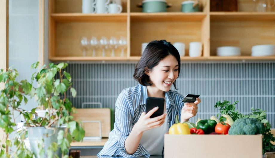 Smiling young woman grocery shopping online with mobile app device on smartphone and making online payment with her credit card, with a box of colourful and fresh organic groceries on the kitchen counter at home