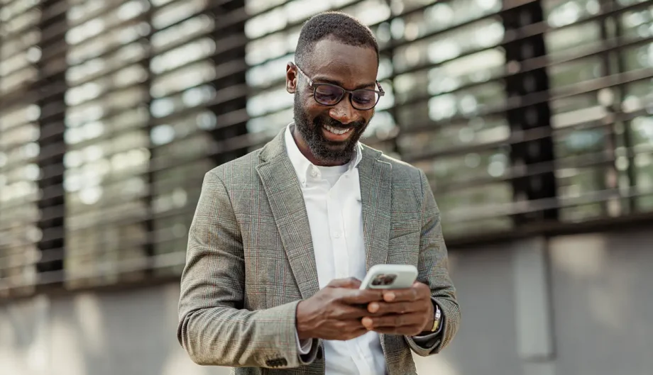Pariveda_Case Study Header_Tech & Digital_Transforming Customer Engagement with AI-Powered Content Optimization A smiling business professional wearing a plaid blazer and glasses stands outdoors in front of a modern building with horizontal slats, looking down at his smartphone.